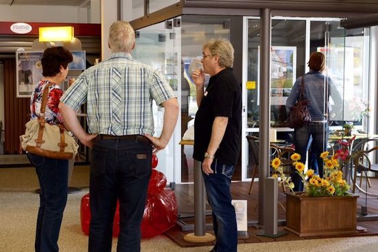 Drei Erwachsene unterhalten sich vor einem Café-Eingang mit Glasfront und Blumendekoration.